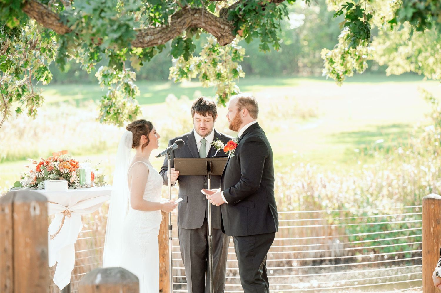An outdoor wedding ceremony takes place under green trees with the couple and officiant at the altar.