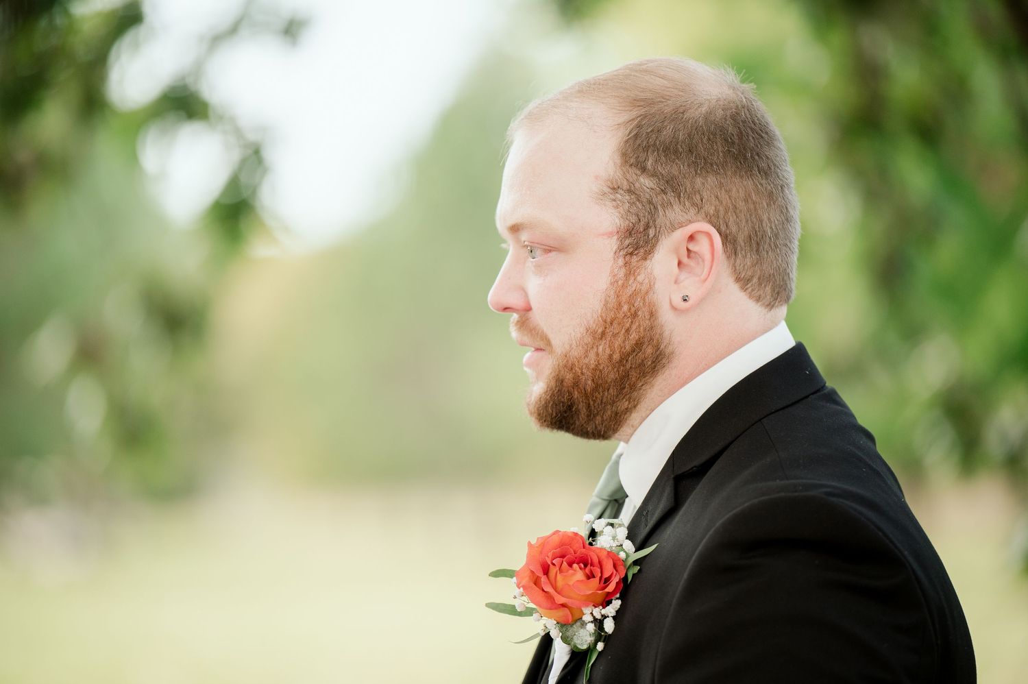 The Grands at Mulligans- Wedding Photos: groom in black suit wearing an orange rose boutonniere against a soft blurred outdoor background.