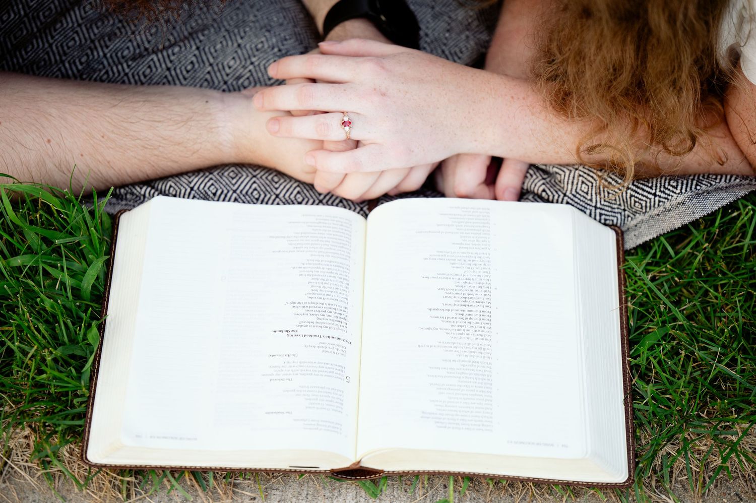 Open book laying on grass with hands resting beside it while someone relaxes outdoors.