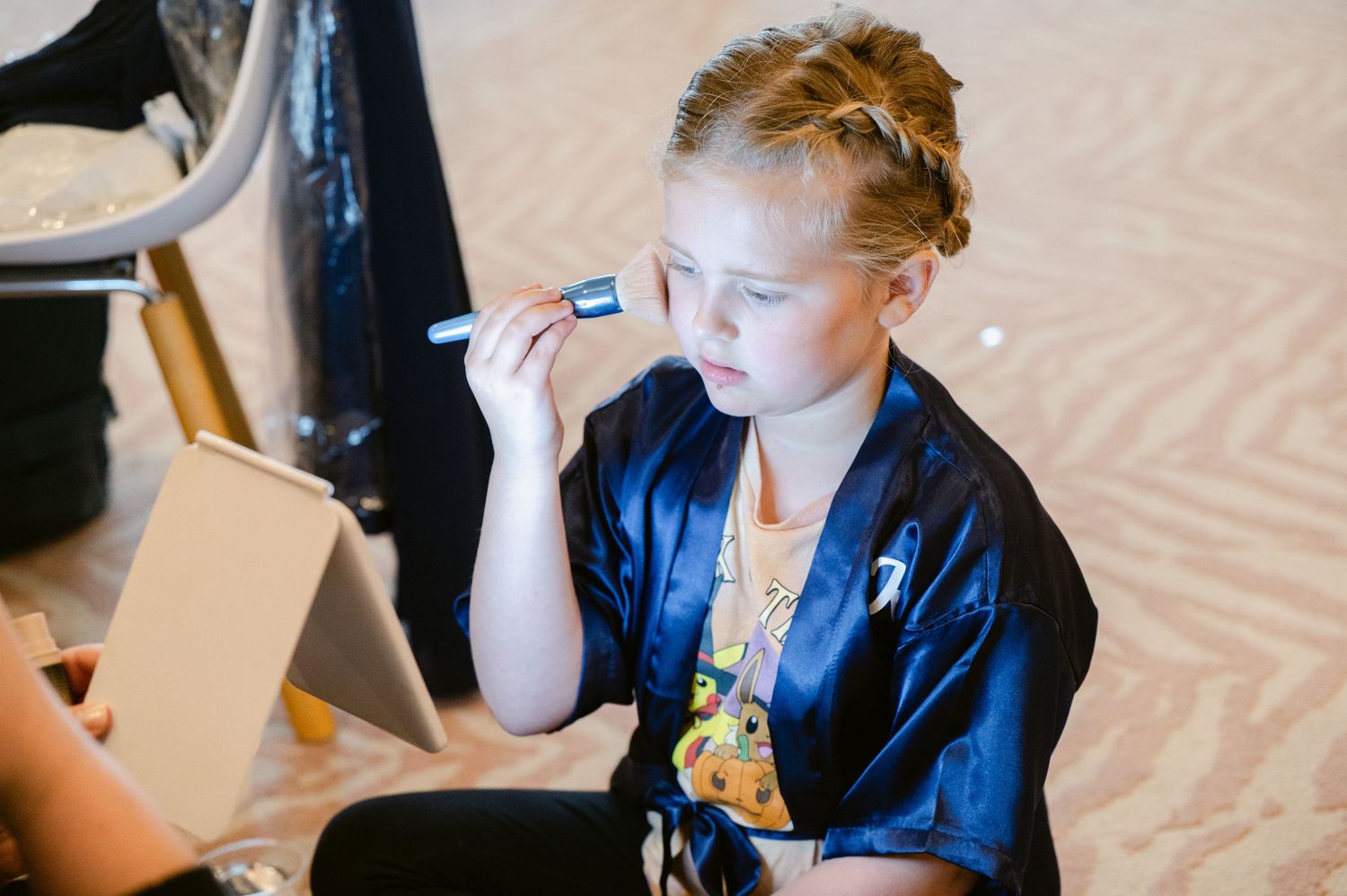 A person in a navy blue robe getting makeup applied while sitting against a cream-colored backdrop.