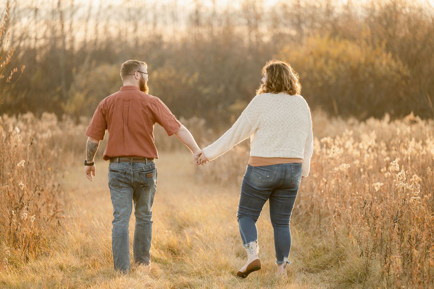A couple holding hands while walking through a golden field at sunset in casual denim and warm clothing. St cloud area engagement photos.