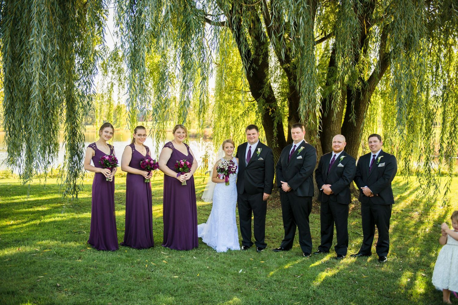 Wedding party stands on lush green lawn under graceful willow trees on a sunny afternoon. Andover wedding at Magestic Oaks Golf Club