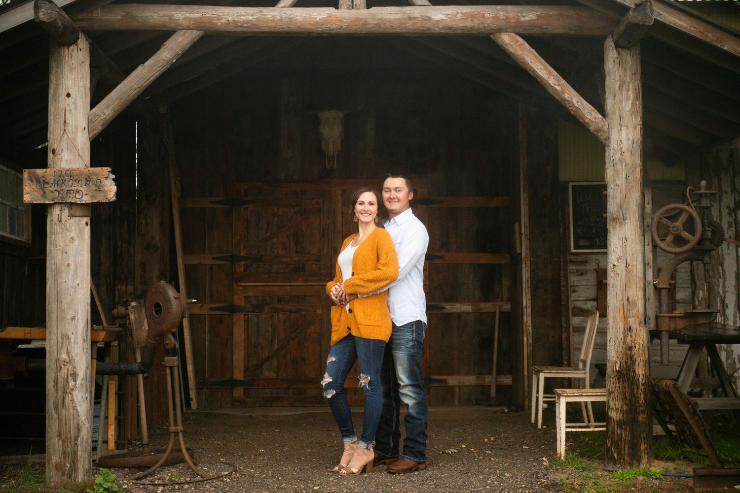 A couple stands together under a rustic wooden barn entrance with vintage farm equipment in the background.