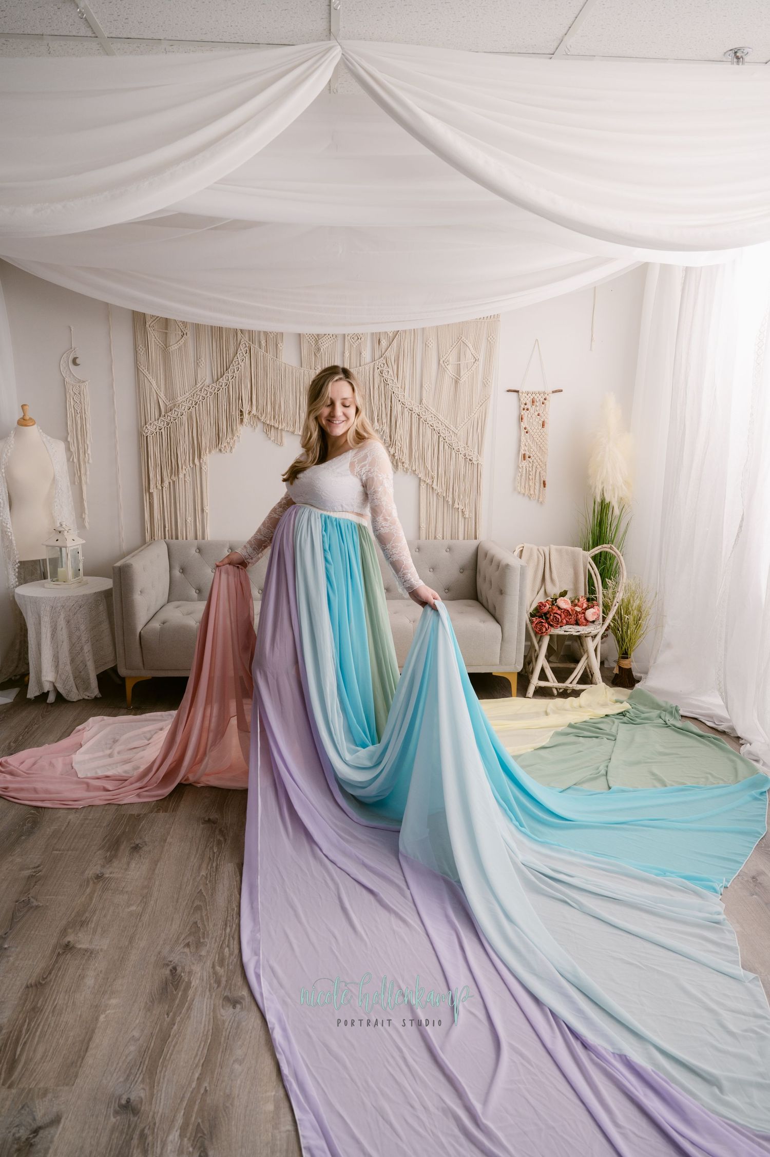 A rainbow colored flowing dress with pastel pink, purple and blue fabric spread out on a wooden floor in a draped white studio.