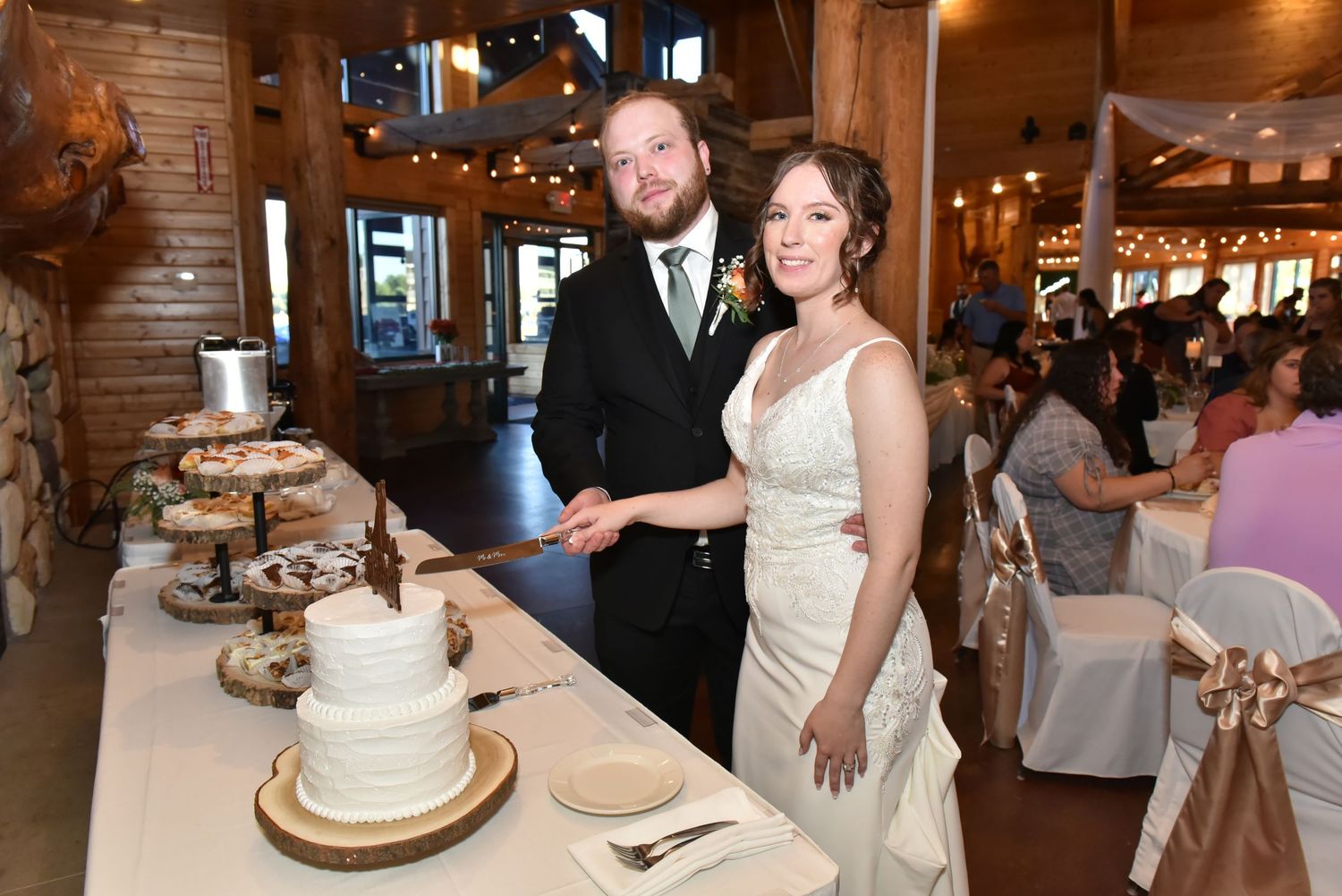 The Grands At Mulligans. A couple cuts their elegant white wedding cake in a rustic wooden reception venue with string lights.