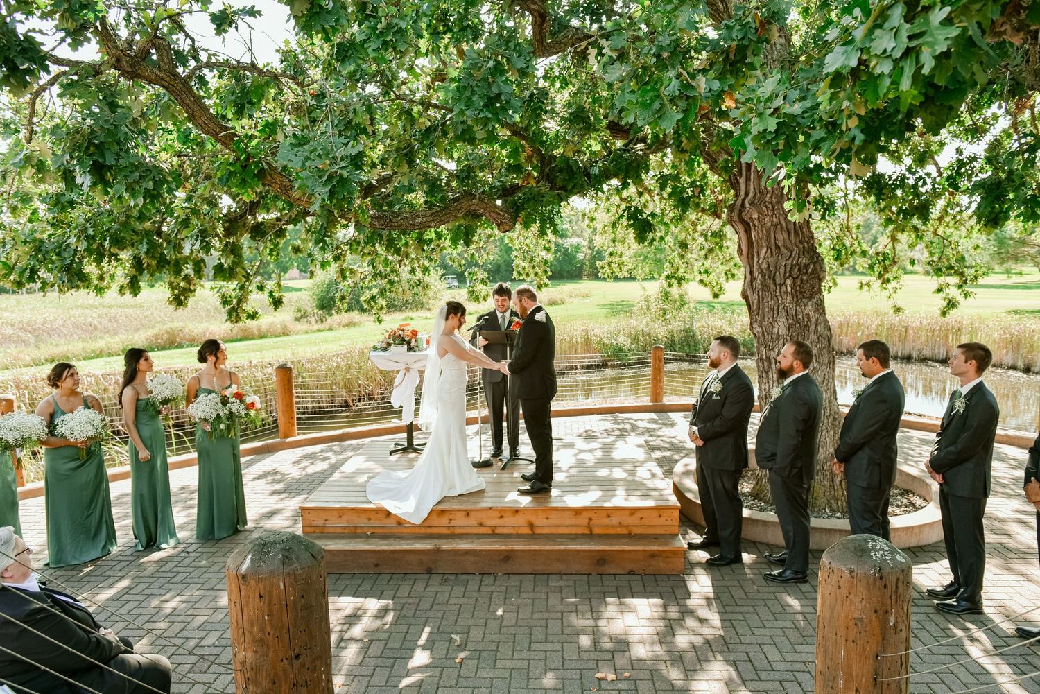 The Grands at Mulligans- Wedding Photos: ceremony under a large tree with wedding party in formal attire on a wooden platform.