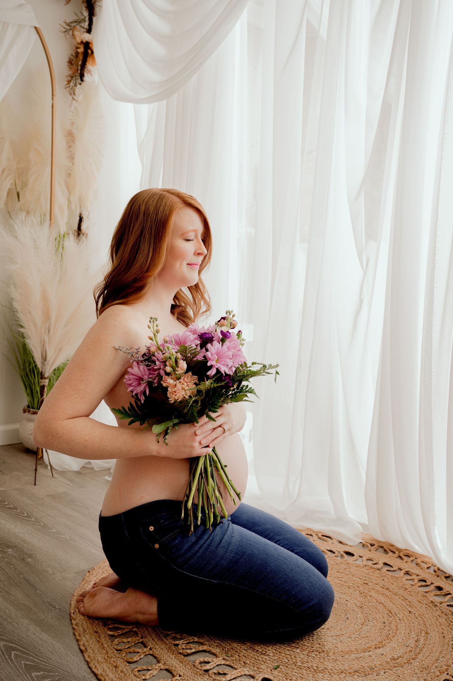 A gentle figure smells a bouquet of pink and purple flowers while sitting peacefully on a round jute rug.