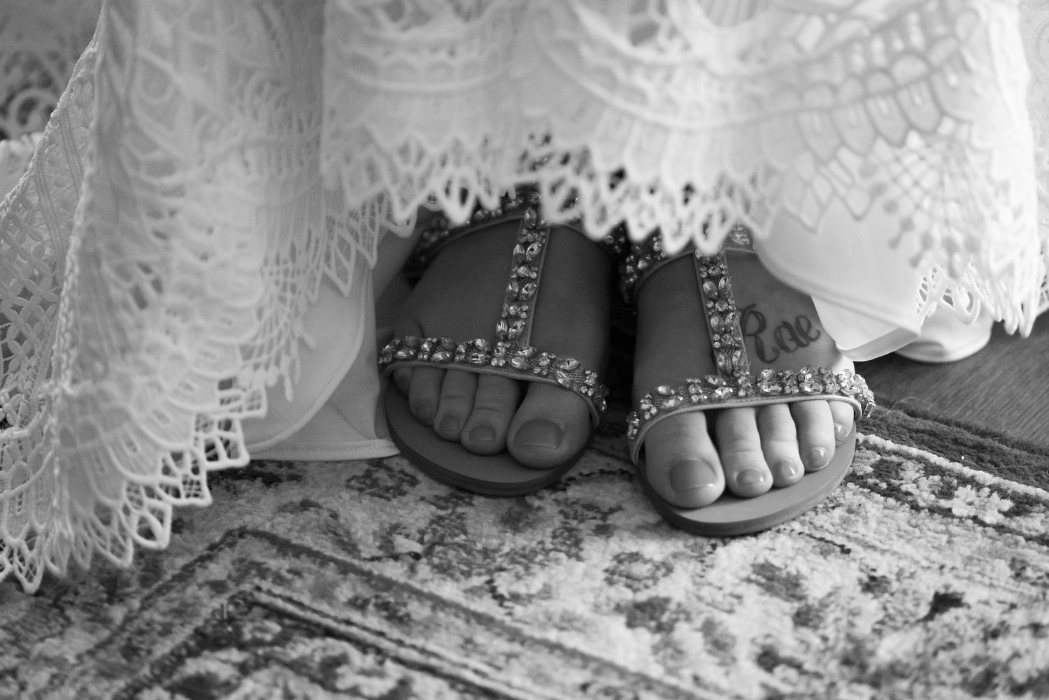 Black and white image of rhinestone-studded sandals peeking out beneath white lace fabric on a patterned rug.