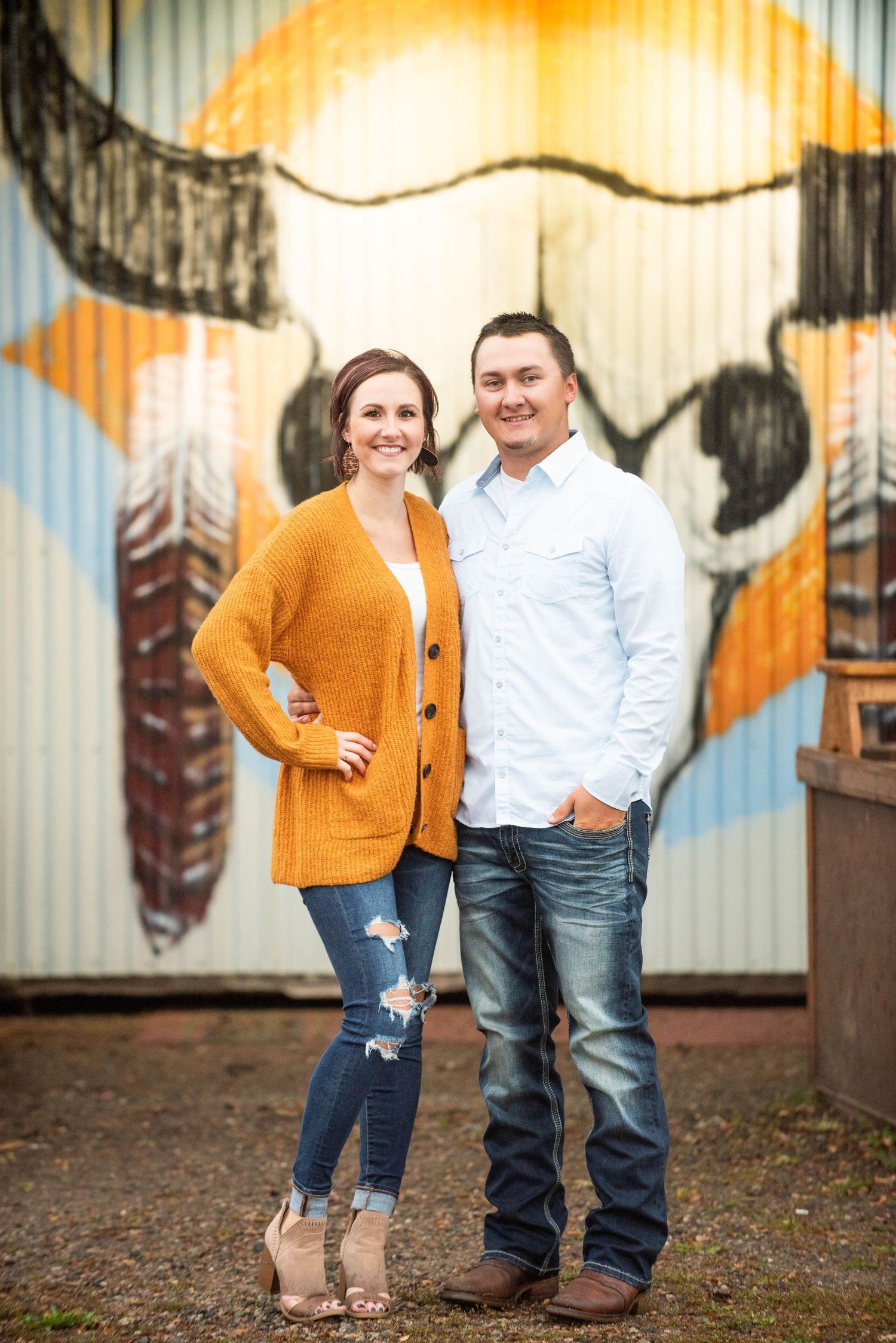 A couple stands together in front of a colorful painted mural wall wearing casual attire.