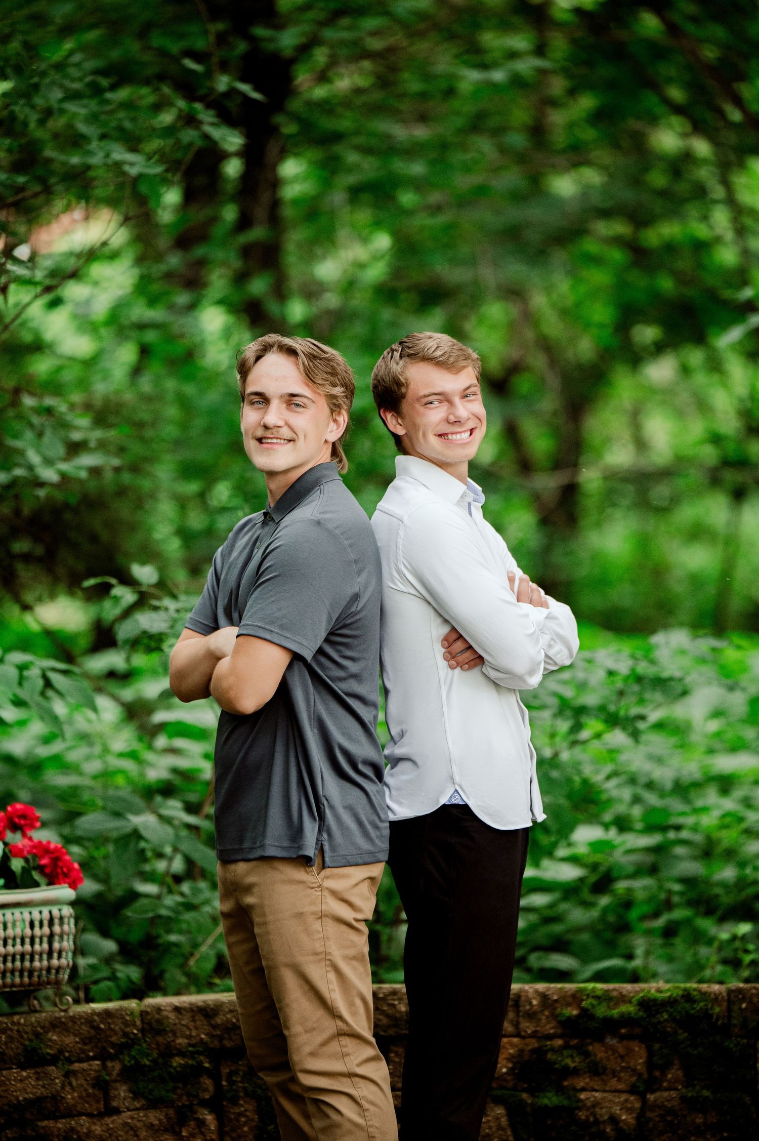 Clear Lake MN Family Photos: Two young people pose back-to-back on a stone wall with red flowers and forest scenery.