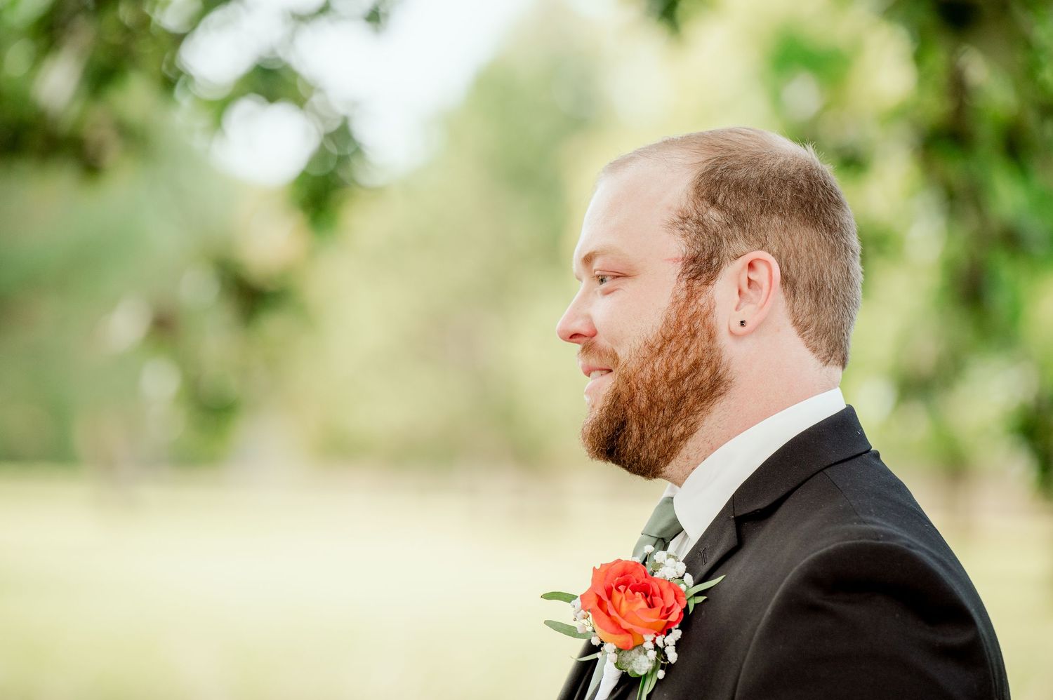 The Grands at Mulligans- Wedding Photos: Close-up of a groom in a black tuxedo with a coral boutonniere in an outdoor garden setting.