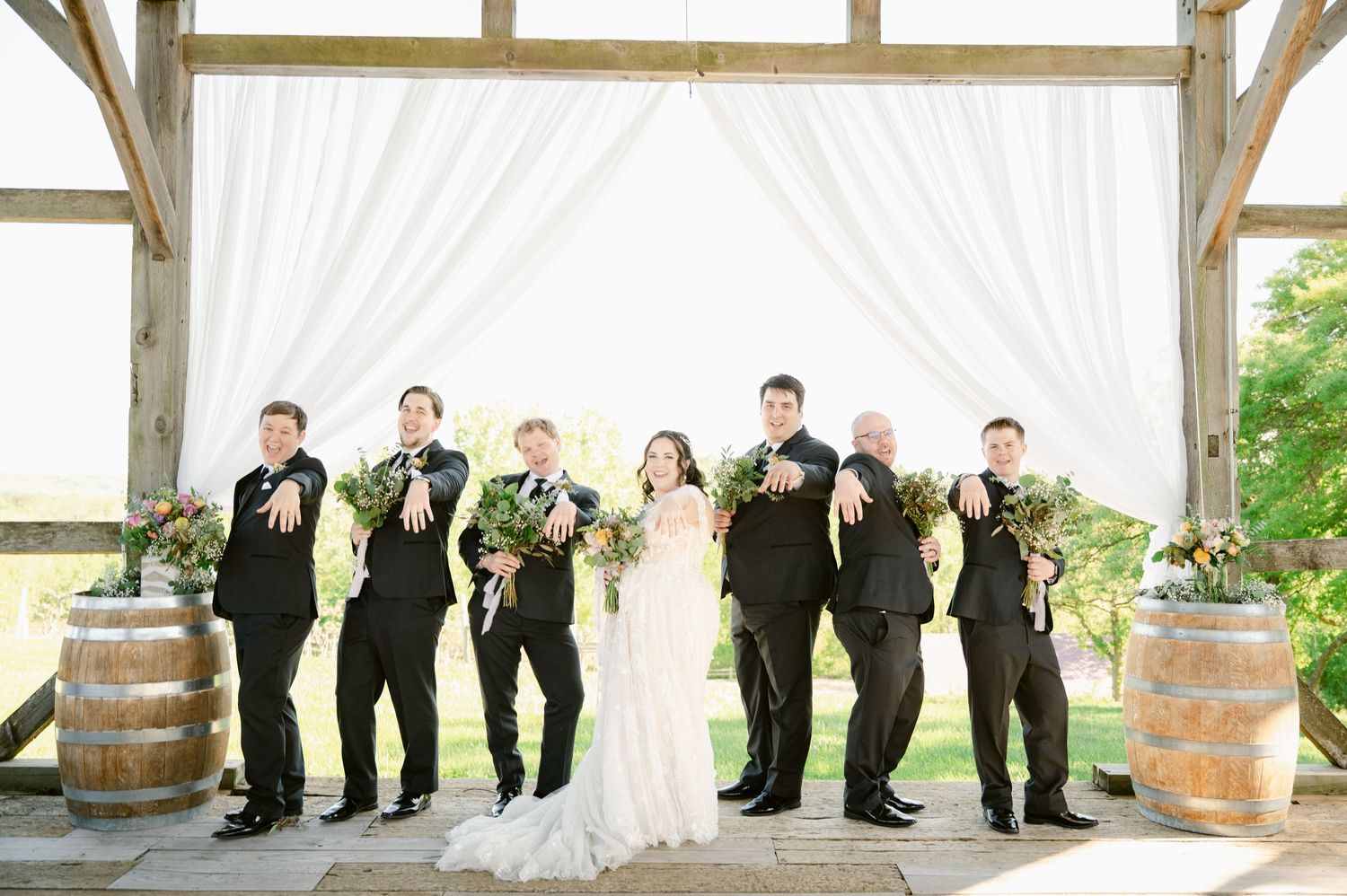 Wedding party in black attire poses playfully under white draped pergola outdoor setting.
