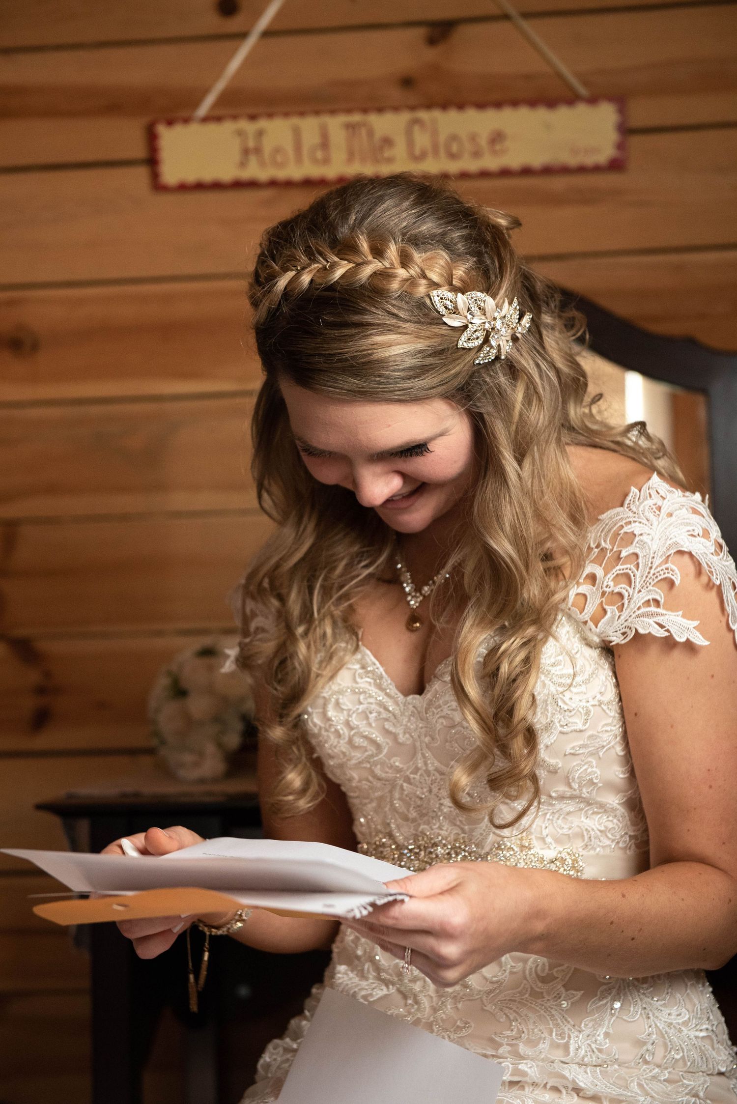 Bride in lace wedding dress with braided hair and crystal headpiece smiles while reading a letter in wooden cabin. Rum River Barn Milaca Min
