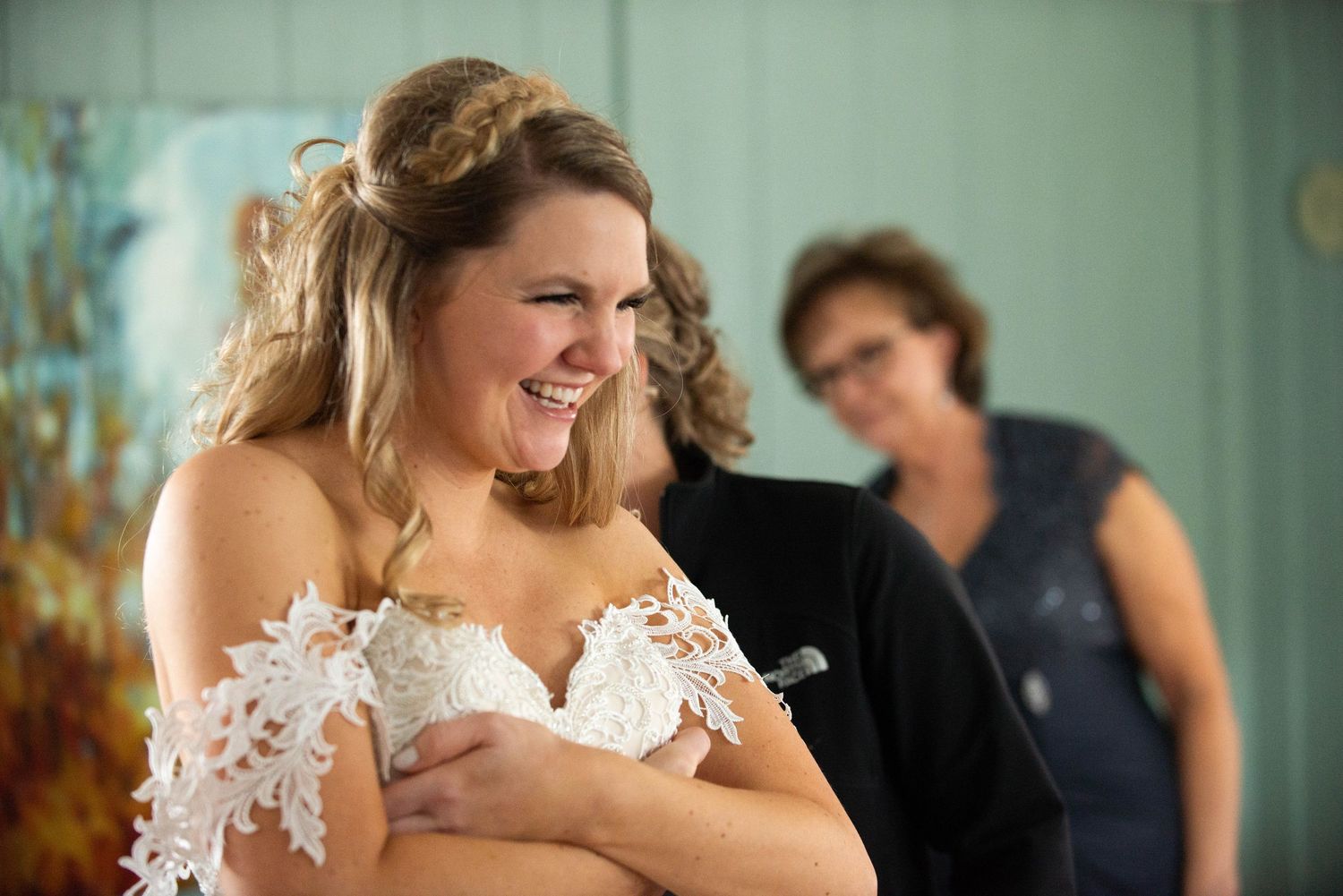 A candid moment catches a bride in a white lace off-shoulder dress laughing joyfully during wedding preparations. Rum River Barn Milaca Minn