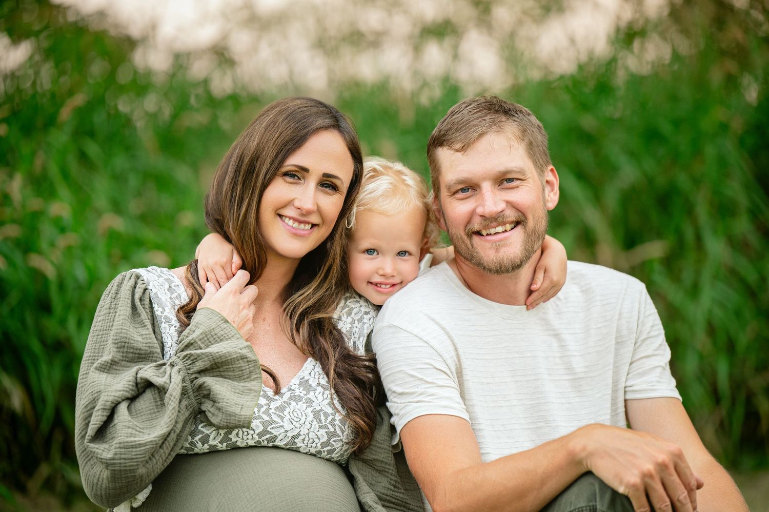 Casual outdoor family portrait showing parents and toddler smiling against natural greenery.