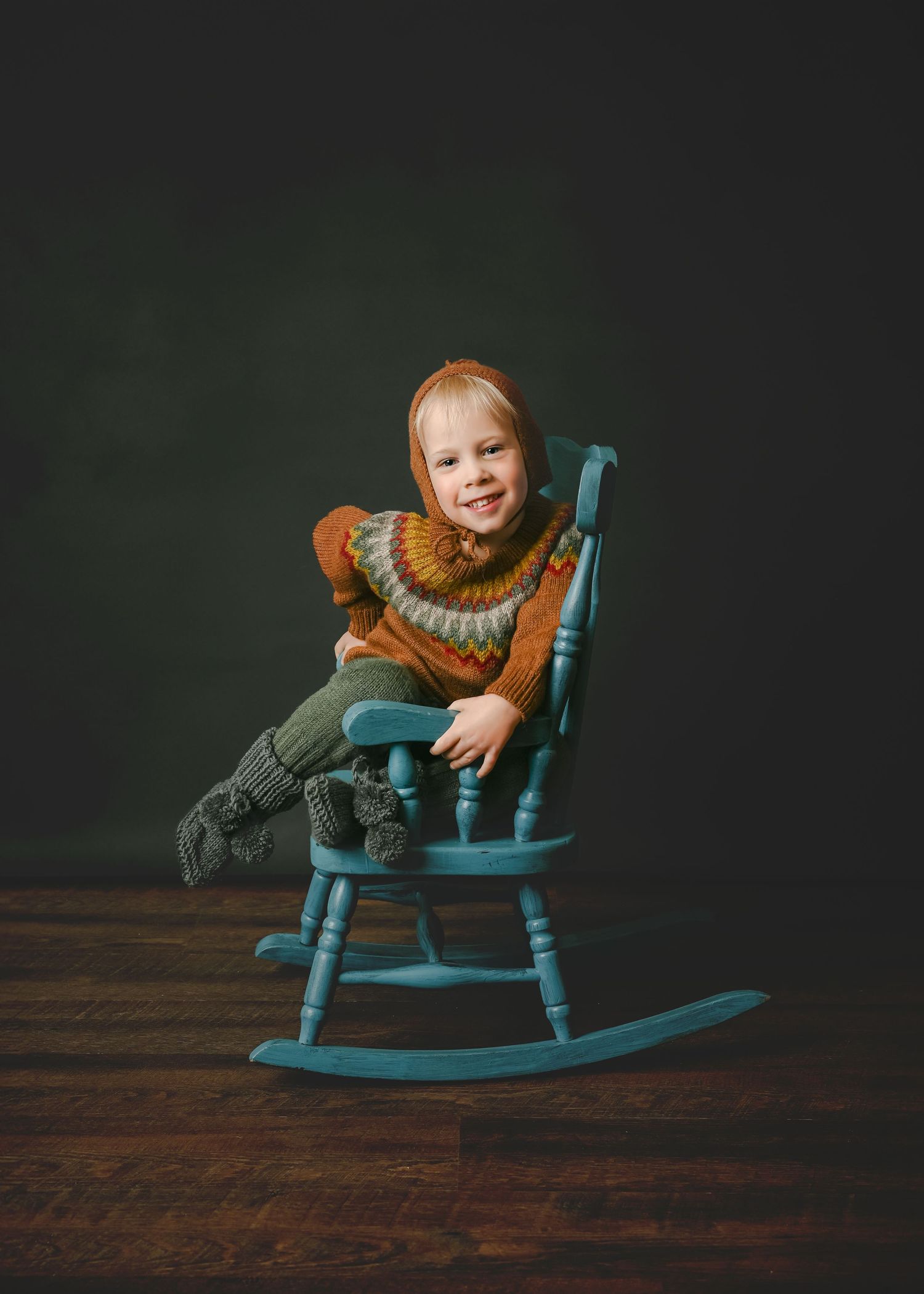 Child wearing orange sweater sits playfully on turquoise rocking chair against dark backdrop.