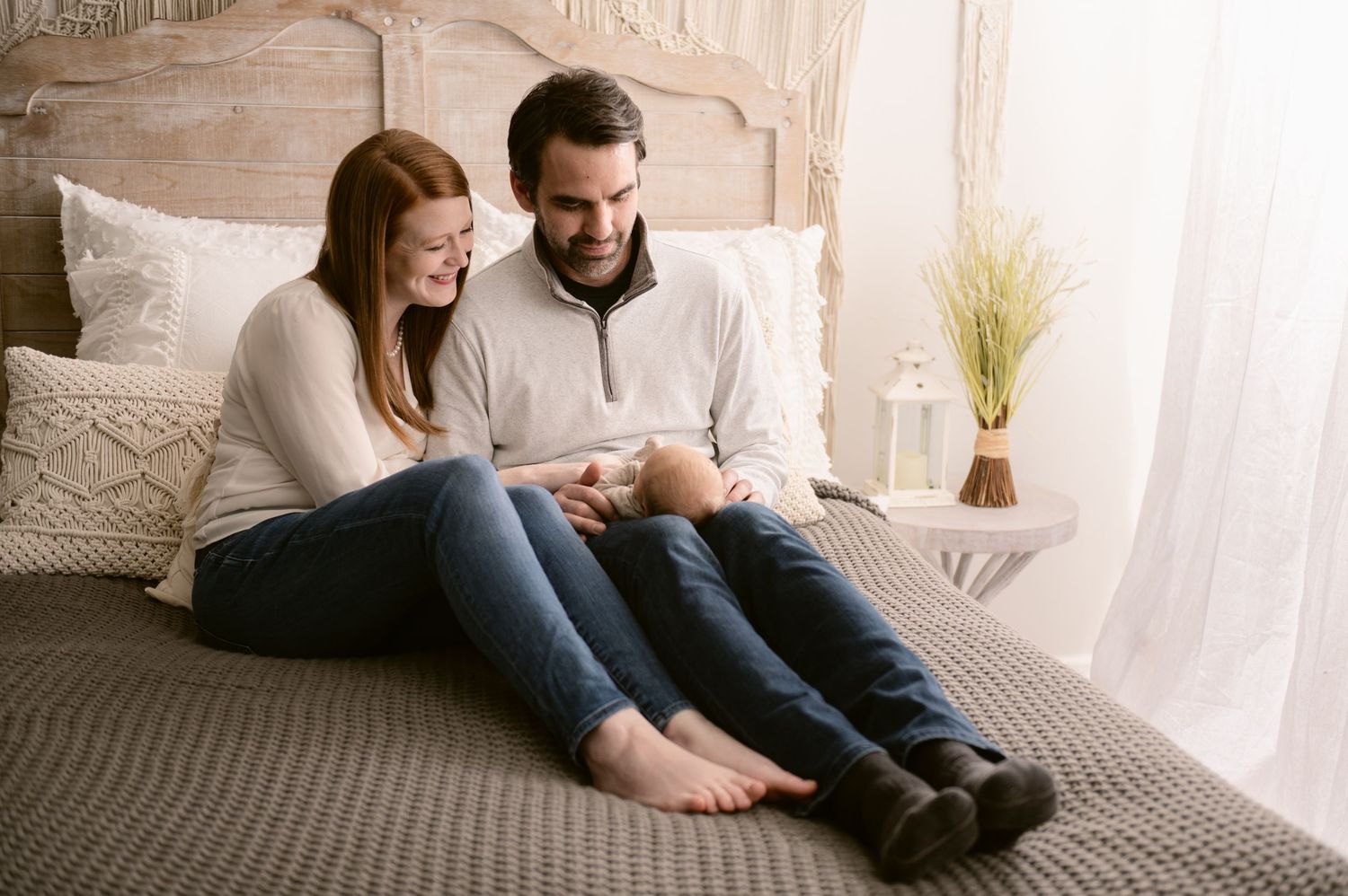 Two people sit together on a grey bedspread in a sunlit bedroom while looking at something between them.