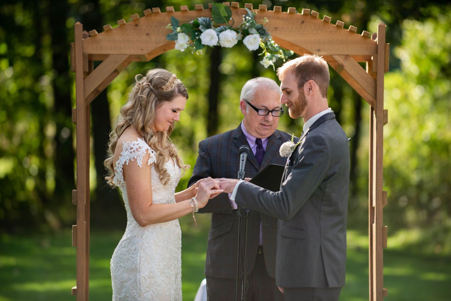 Wedding ceremony under a wooden arch with exchange of rings in a garden setting. Rum River Barn Milaca Minnesota Weddings