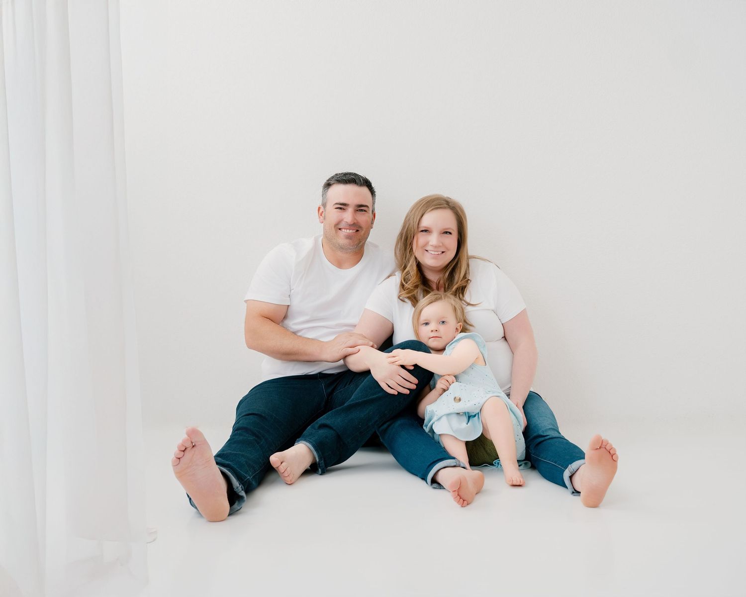 A family wearing white and denim sits together against a white backdrop for a casual portrait session.