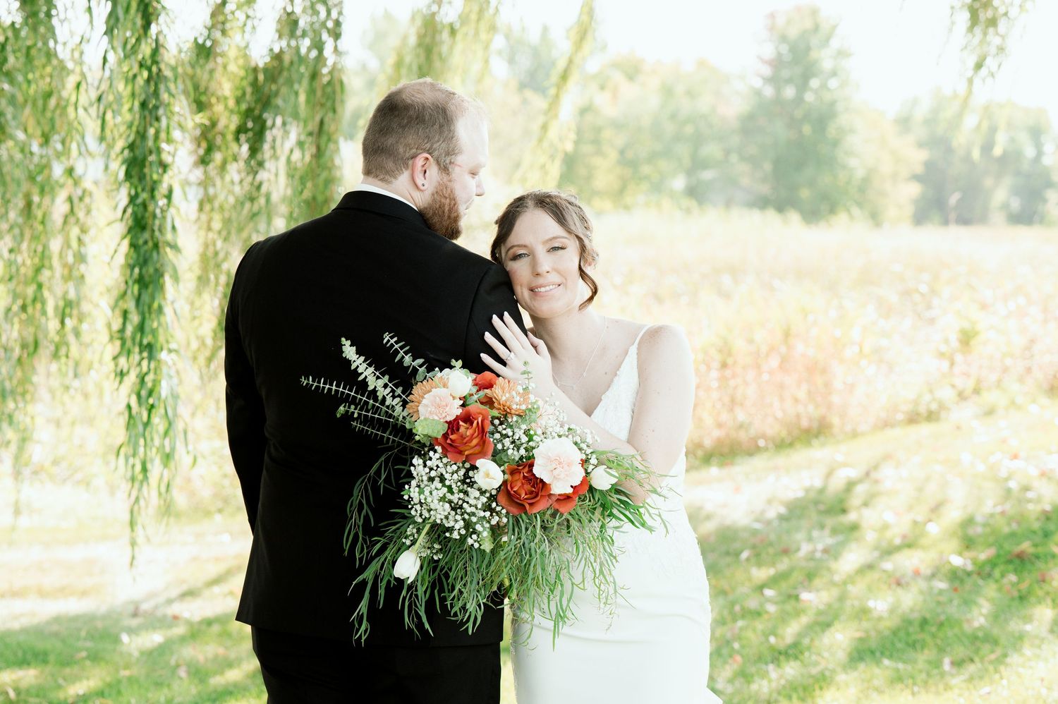 Wedding couple embraces under weeping willow trees in a sunlit garden setting.
