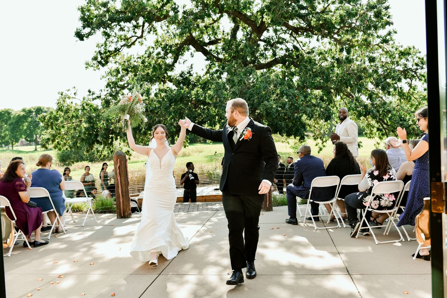 The Grands At Mulligans Bride and groom coming up the aisle. Wedding guests seated in white chairs under tree shade.