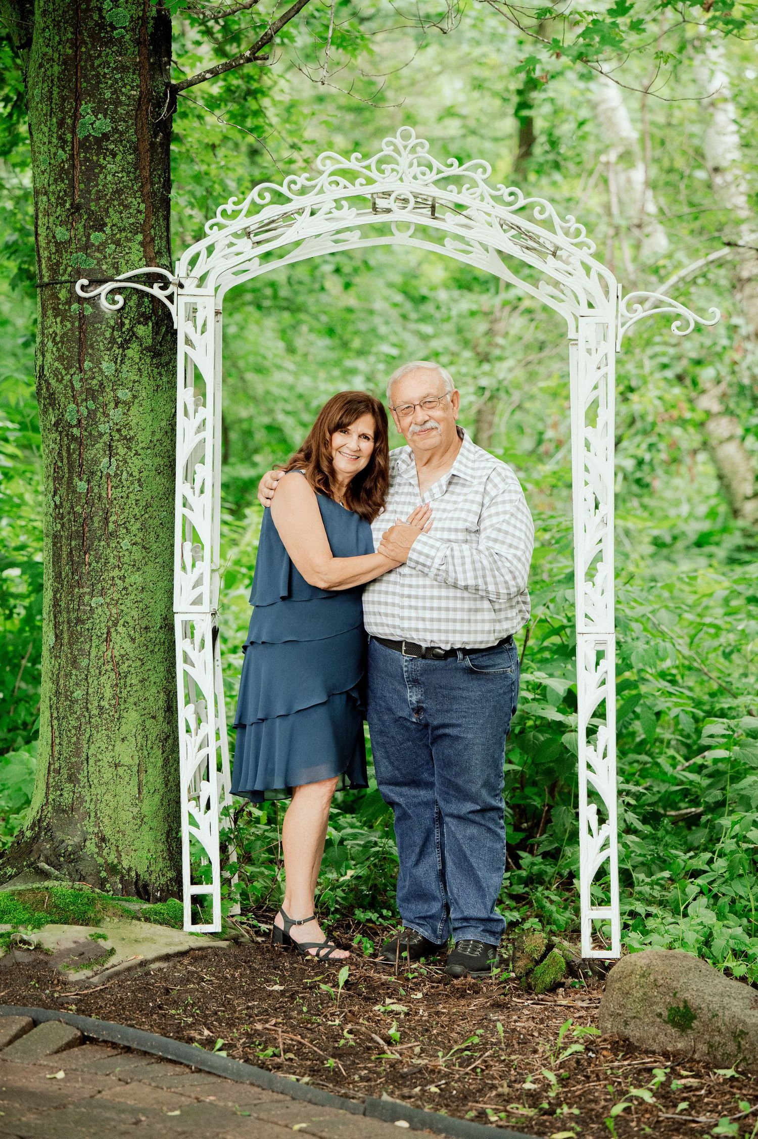 Clear Lake Family Photography: A couple poses together under an ornate white metal garden arch against a verdant forest backdrop.