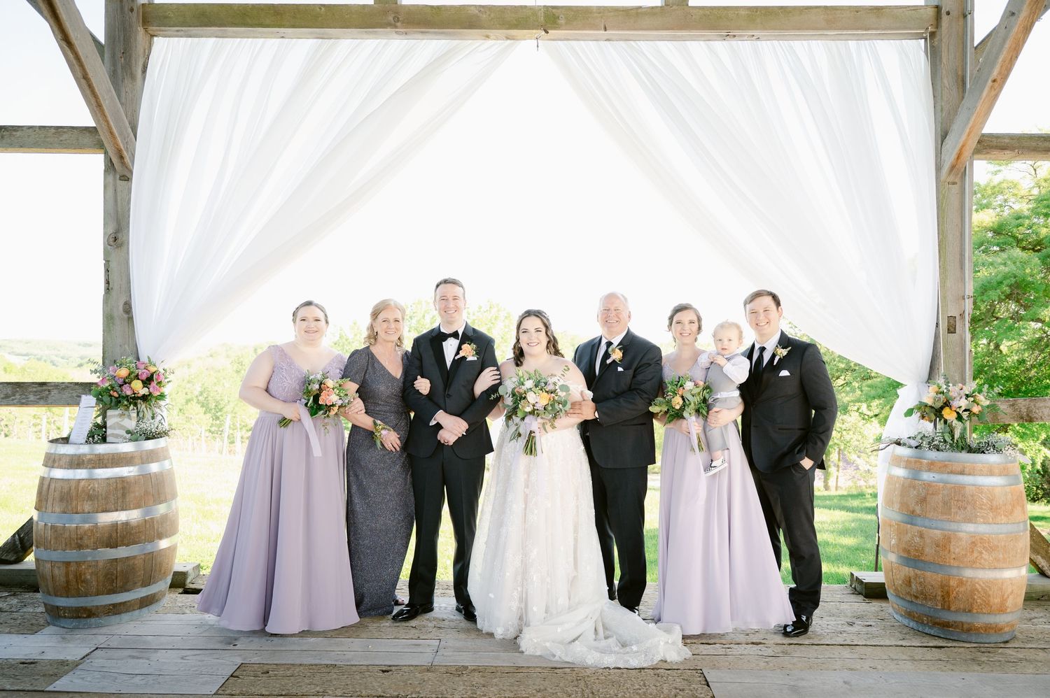 Wedding party poses under white draped pavilion with rustic wine barrels.