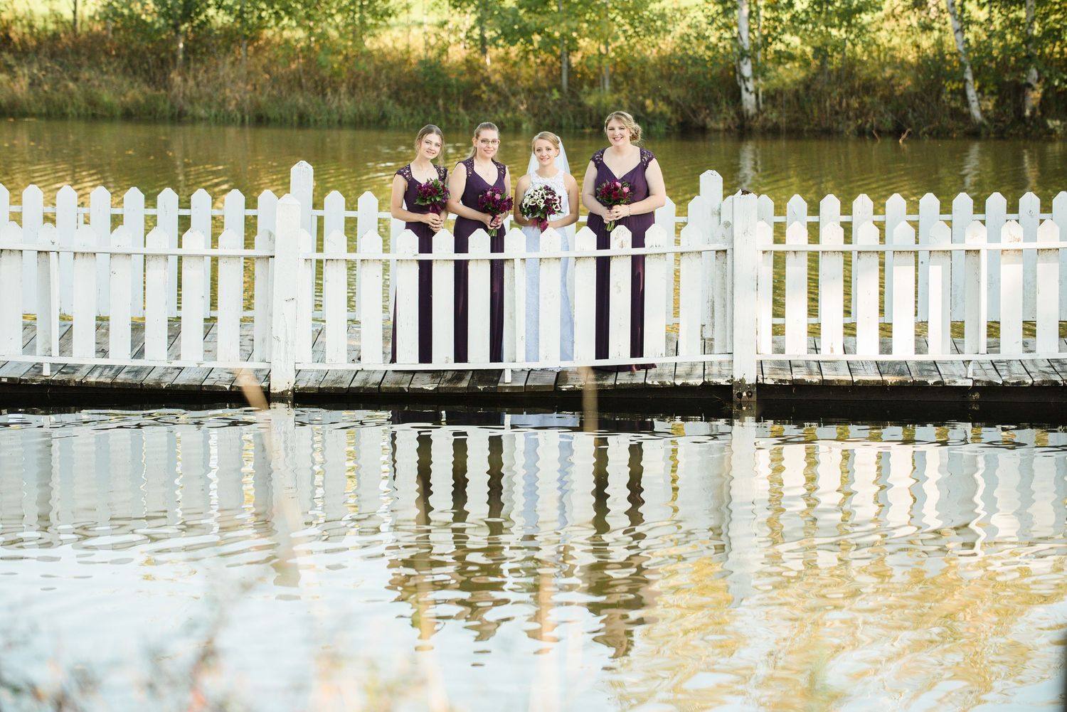 Bridesmaids in dark dresses pose on a white bridge over water during sunset, creating beautiful reflections. Andover wedding at Magestic Oak