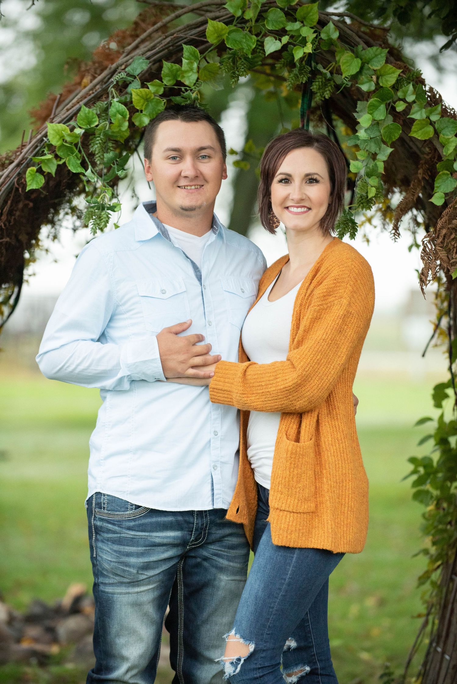 A couple shares tender moments under green foliage in a park setting wearing casual outfits of blue and orange.