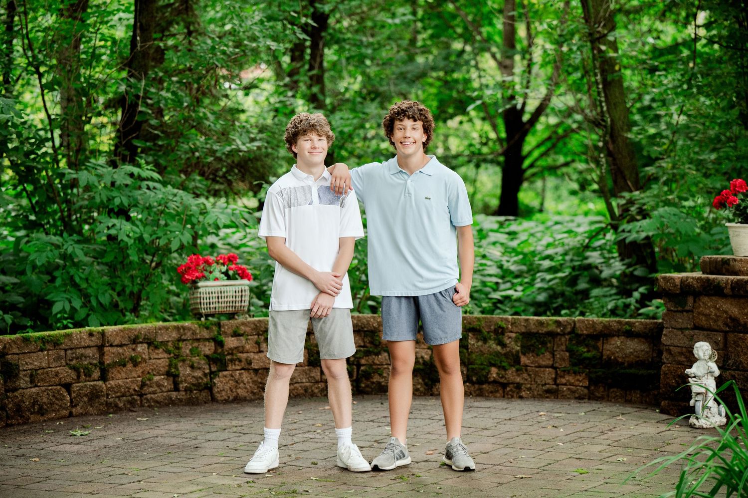 Clear Lake MN Family Photos: Two people standing on a stone patio surrounded by lush green trees and red flowers in a garden setting.