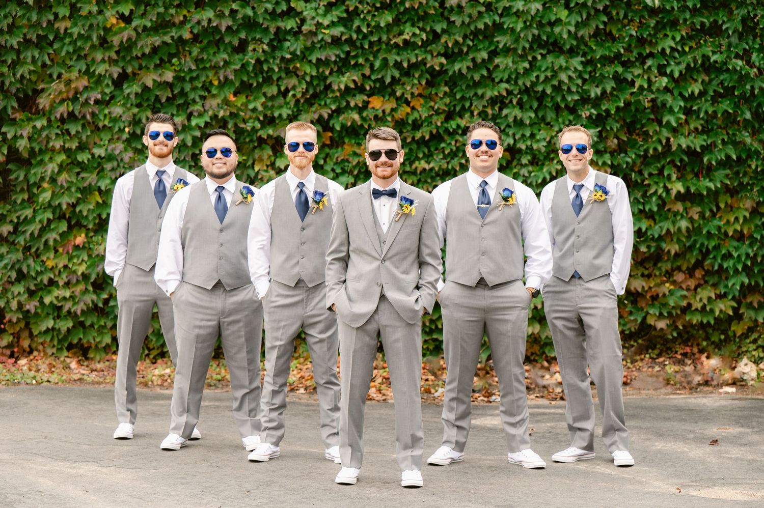 Wedding party in matching grey suits and sunglasses pose against an ivy covered wall.