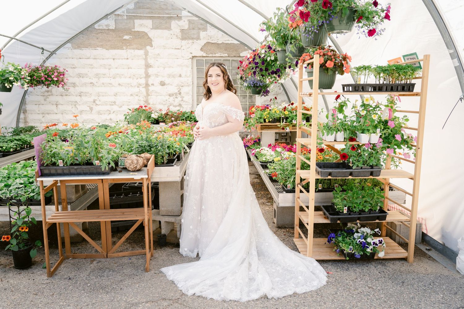 A wedding dress displayed in a greenhouse setting surrounded by colorful hanging flowers and wooden shelves.