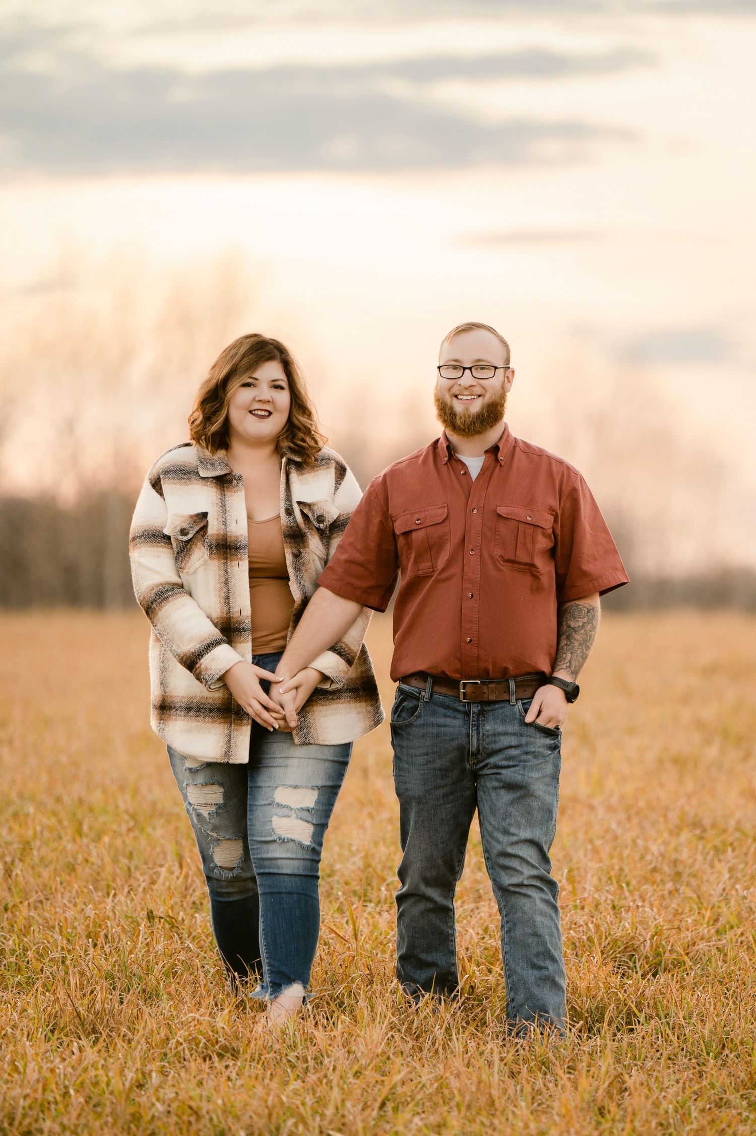 A couple stands together in a sunlit golden field at sunset wearing coordinating denim and brown outfits. St cloud area engagement photos.