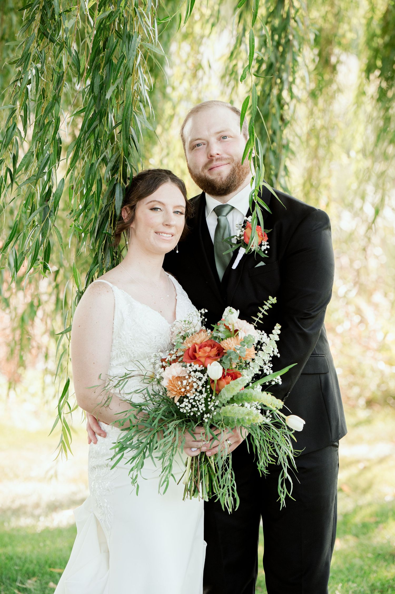 A couple poses under a weeping willow tree with a colorful autumn bouquet featuring orange and white flowers.