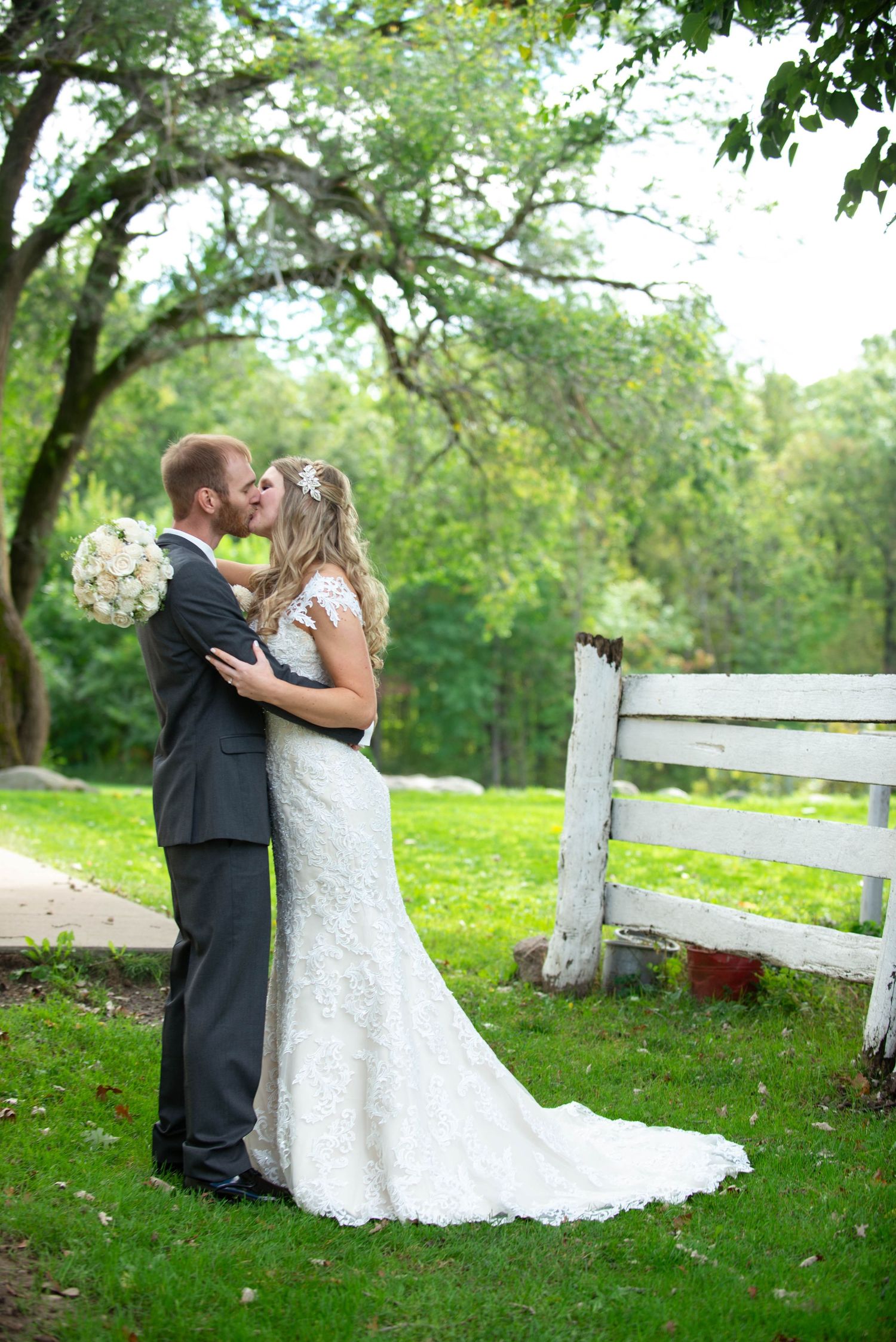 A wedding couple shares a romantic moment by a white fence in a lush green outdoor setting on their special day. Rum River Barn Milaca Minne