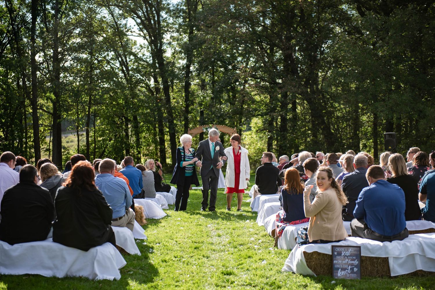 Sunny outdoor wedding ceremony in progress with guests seated on white chairs in wooded setting. Rum River Barn Milaca Minnesota Weddings