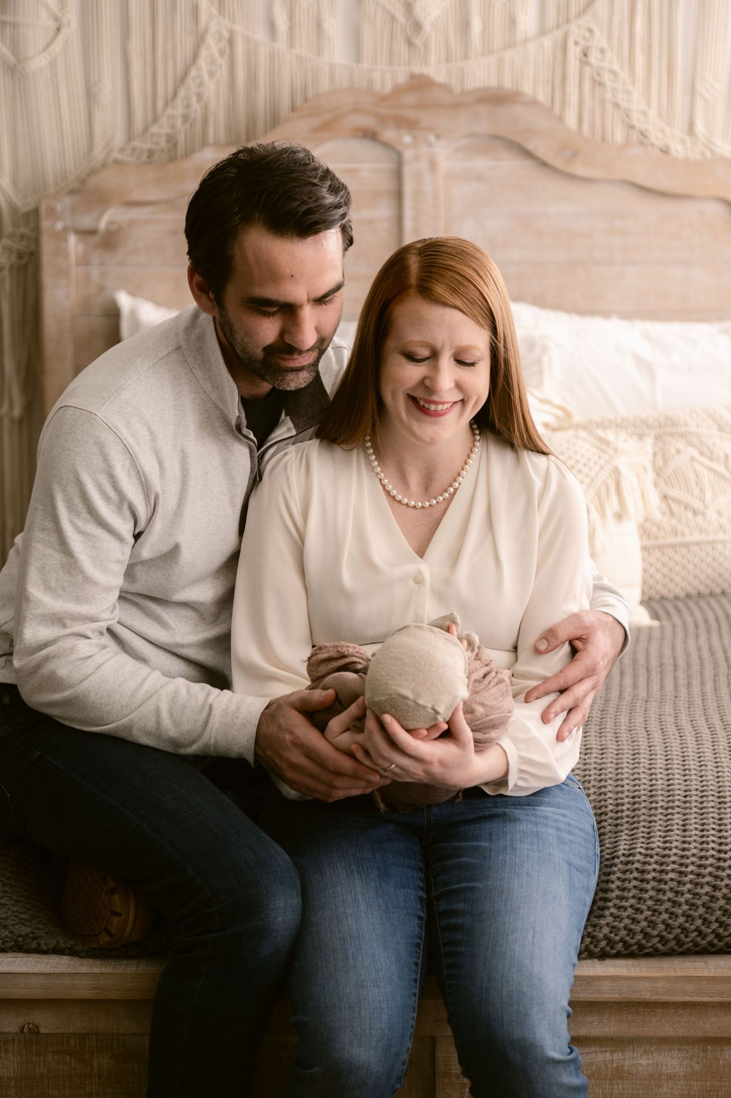 A couple sits together on a bed as they lovingly cradle their newborn baby wrapped in beige fabric.