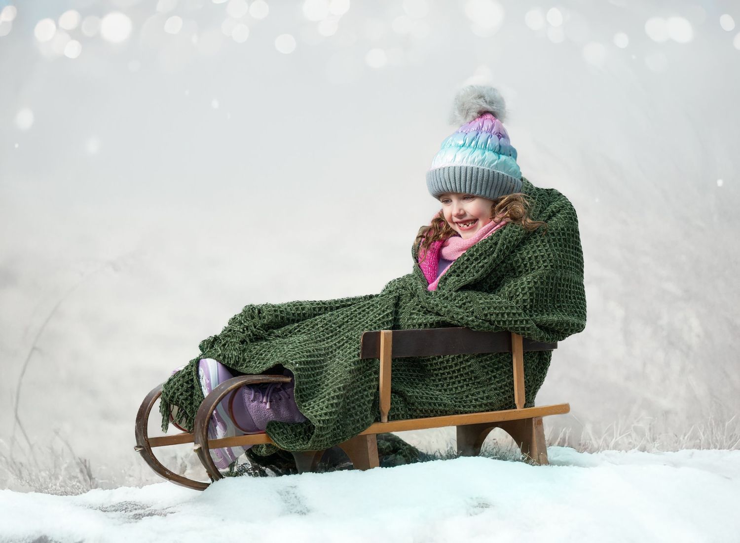 A winter scene shows someone bundled up in a cozy knit hat and sweater sitting on a vintage wooden sled in the snow.