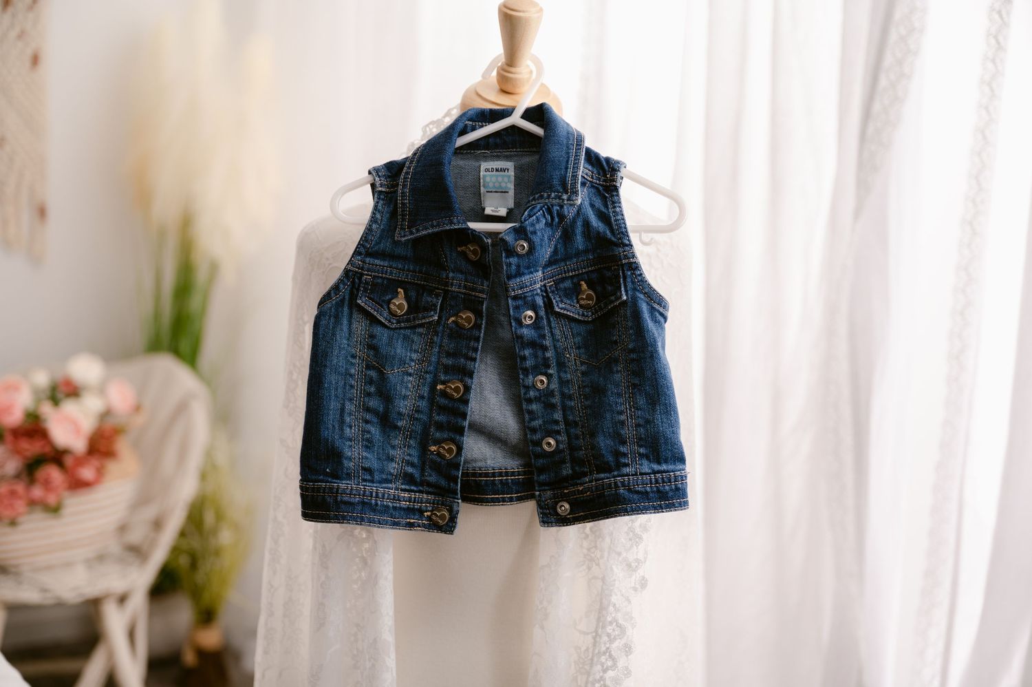 A sleeveless blue denim jean vest hangs against a white curtain background with pink flowers visible.