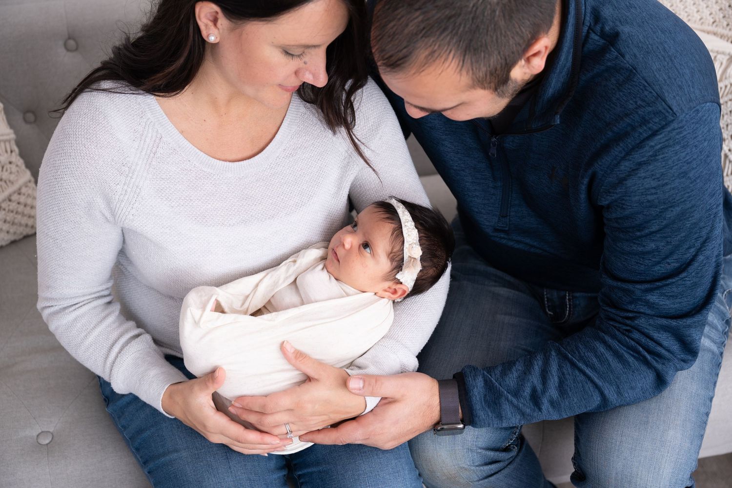 Newborn baby swaddled in white being held lovingly by parents on gray couch.
