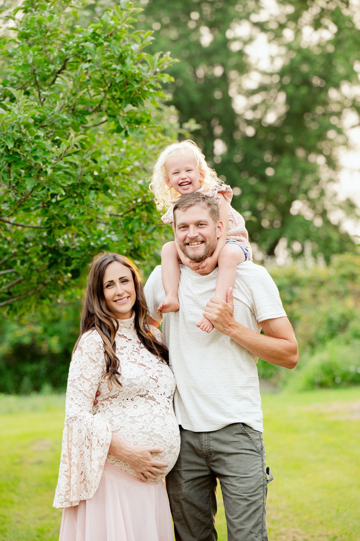Expectant parents and toddler pose for a family portrait in a lush green outdoor setting.