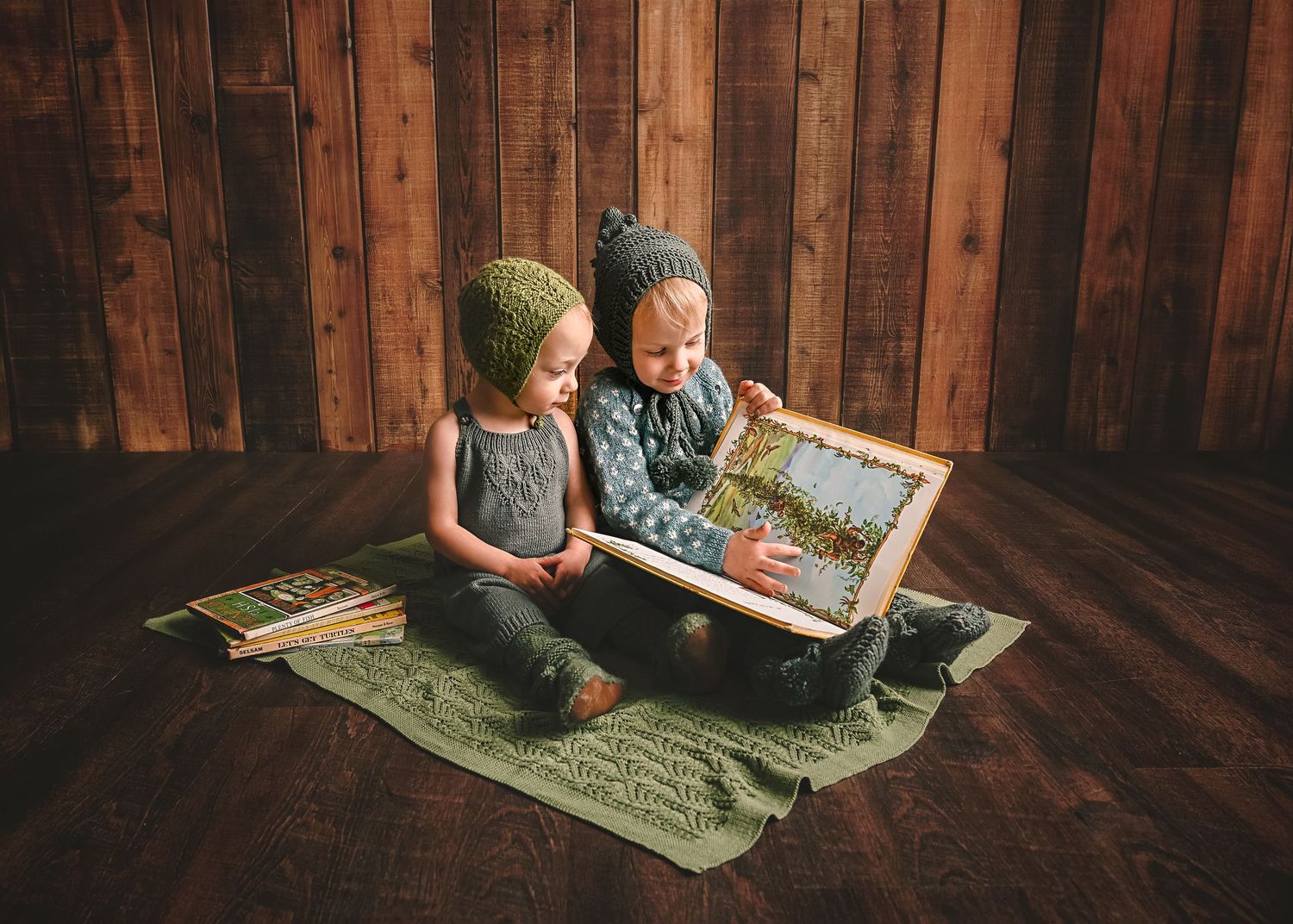 Two small ones reading picture book together while sitting on blanket against wood wall.