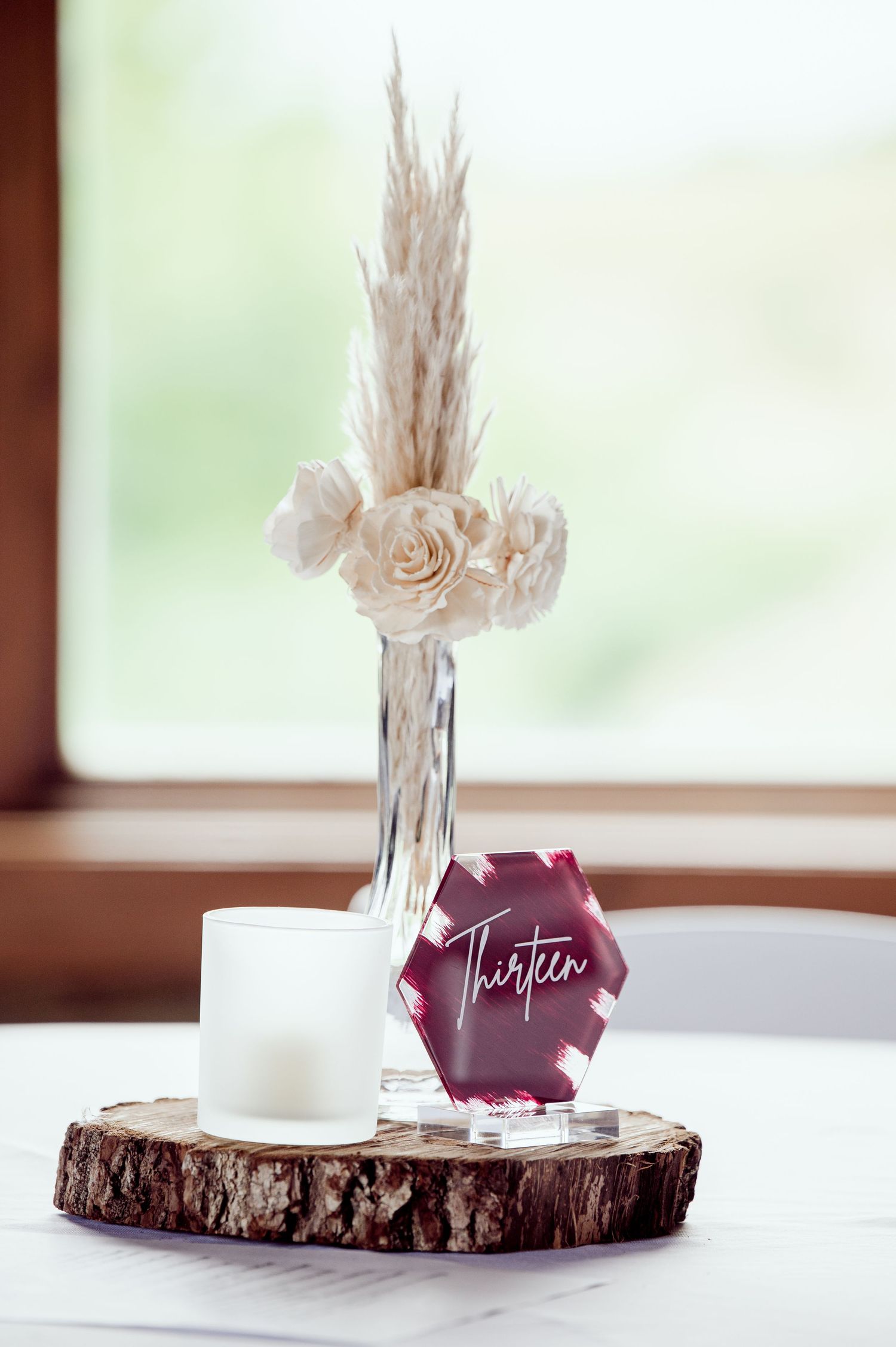 Wedding centerpiece featuring dried pampas grass and roses on a wooden slice with burgundy accents.