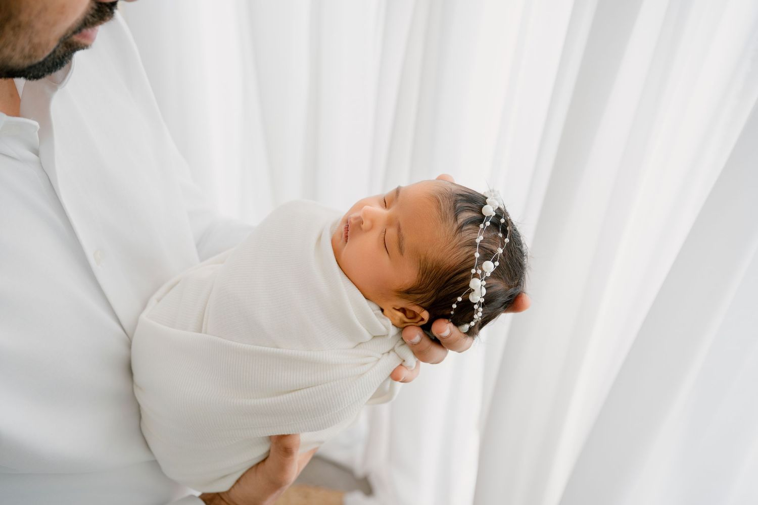 Parent holds sleeping swaddled newborn wearing delicate crown against white backdrop.
