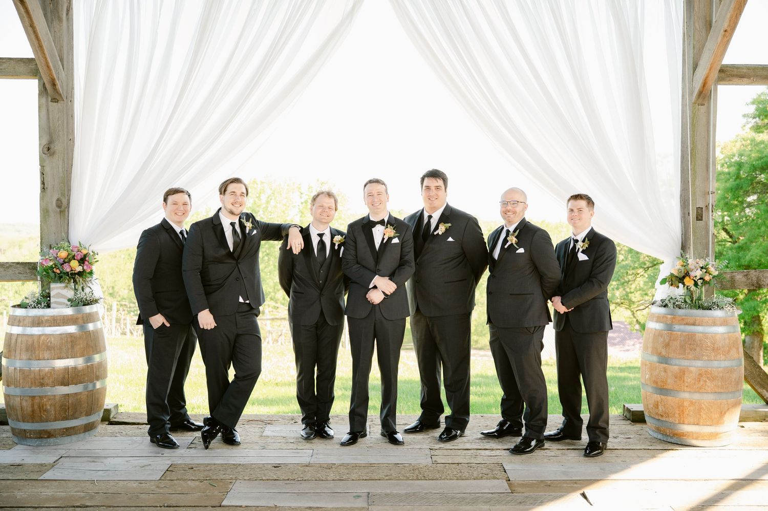 Wedding party in black tuxedos poses under white draped pergola with rustic wine barrel decor.