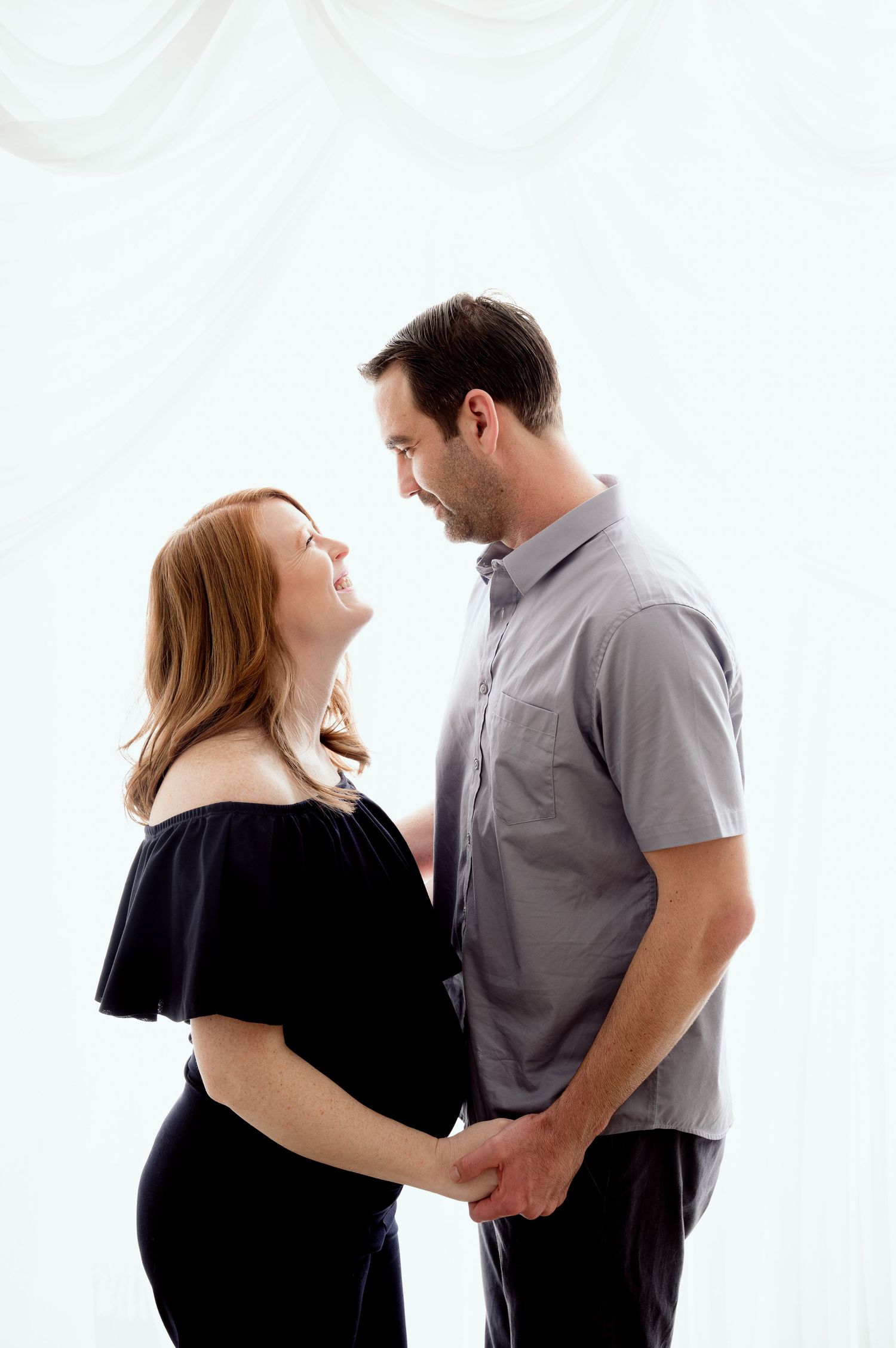 Couple in casual attire sharing a tender moment against white backdrop during maternity photoshoot.