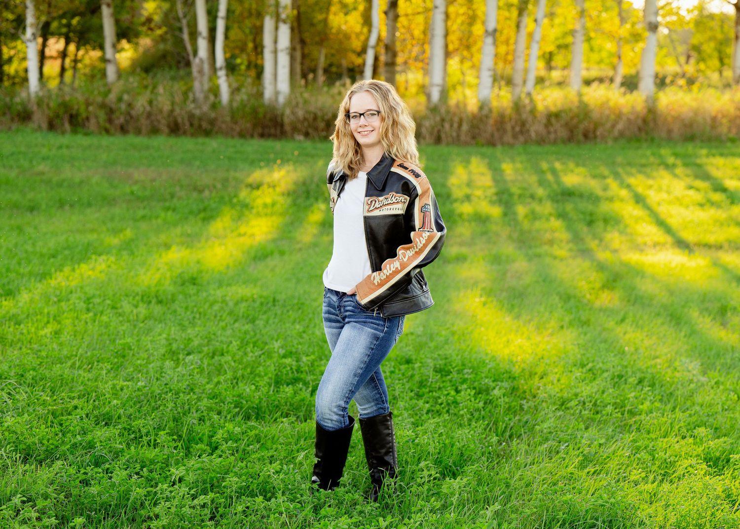 Senior Portrait in field with tall boots against backdrop of golden autumn trees and sunlit grass.