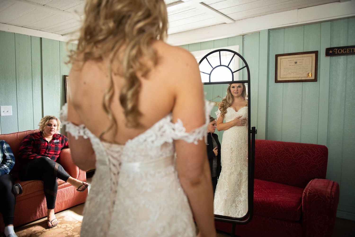 A detailed backside view of an ivory lace wedding gown being fitted in a mint green room with a mirror and red couch.