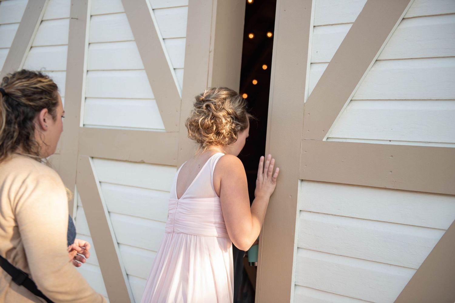 A young person in a white dress peers through a wooden door frame decorated with twinkling lights. Rum River Barn Milaca Minnesota Weddings