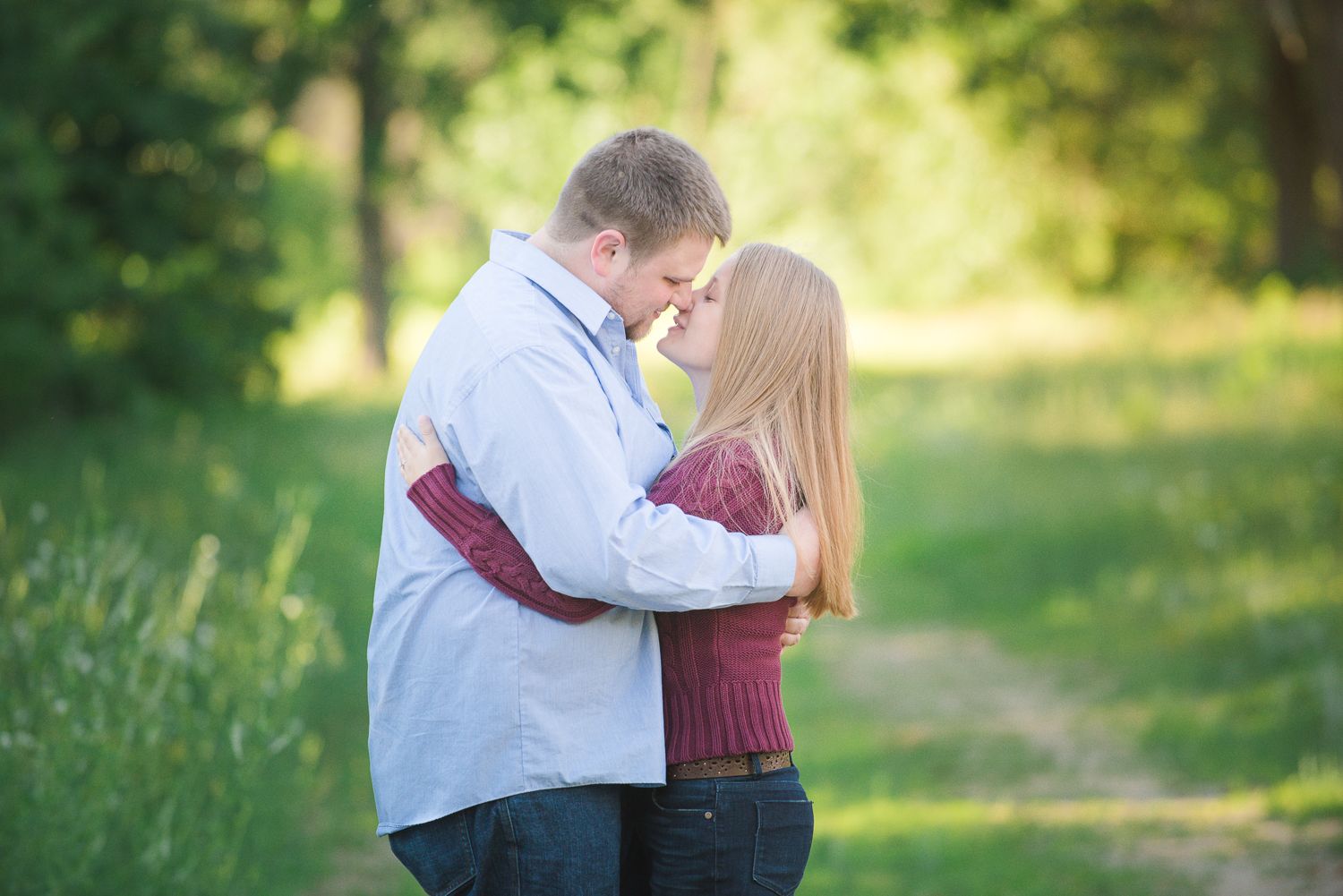 A tender moment between a couple embracing on a sunlit path with green grass and trees in the background.